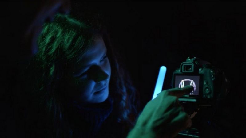 Dans l'obscurité, une femme tenant un bâton lumineux regarde l'écran d'un appareil photo Canon tandis qu'une autre personne pointe son doigt vers l'écran.