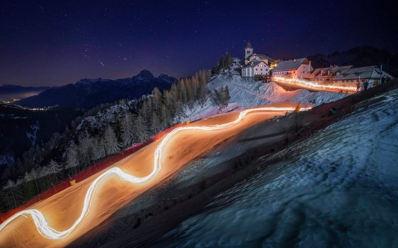 שובלי אור בלילה בכביש שמתפתל על צלע גבעה מושלגת ומיוערת לעבר קבוצה של בניינים לבנים עם גגות אפורים.