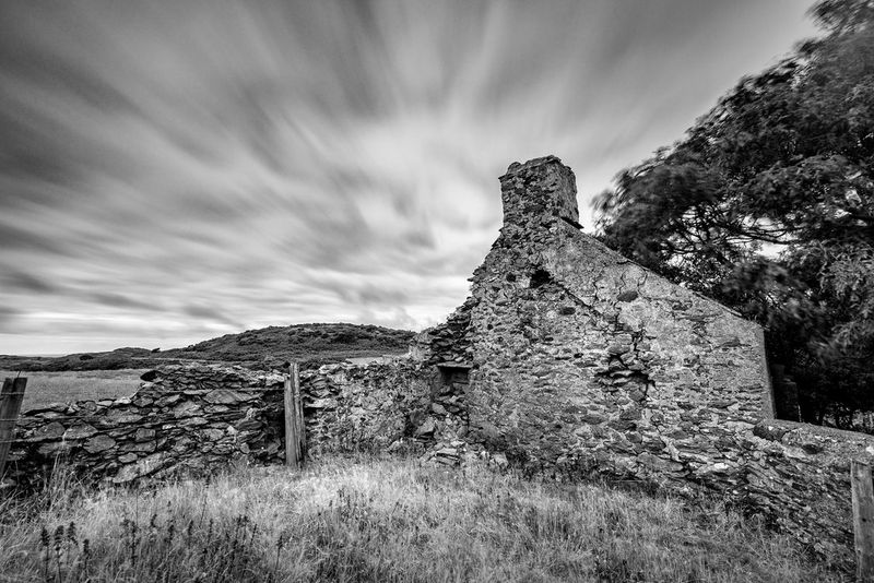 Le rovine di un'antica fattoria, con alberi e un campo sullo sfondo e sopra di essi nuvole catturate in movimento.