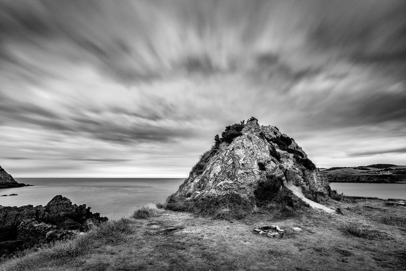 Un tumulo di terra sul bordo di una scogliera, con un mare calmo sullo sfondo e sopra nuvole catturate in movimento.