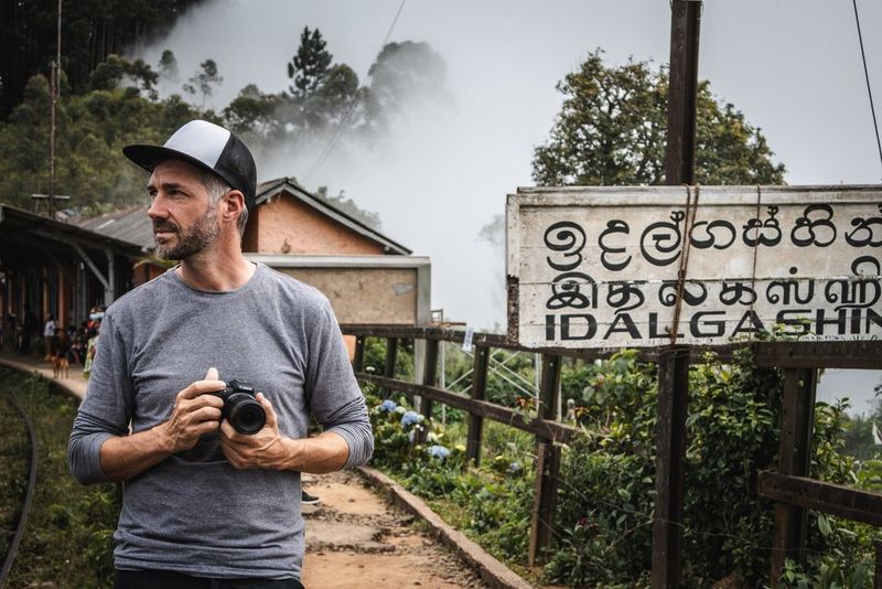 Photographer Martin Bissig holds a Canon EOS R10 camera next to a sign outside a Sri Lankan train station.
