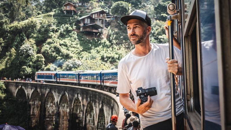 Photographer Martin Bissig leans out of a train that has stopped to pick up passengers on a viaduct in Sri Lanka. In his hand is a Canon EOS R10 and behind him a hill with small wooden houses built into it. Photographed by Monika Bissig on a Canon EOS R7. 