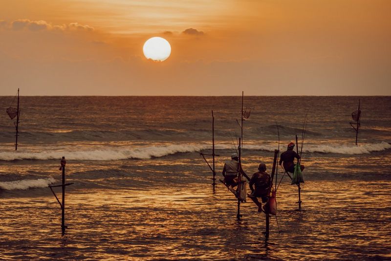 At dusk, three fishermen balanced on poles in shallow water wait patiently for a catch as the sun sets in front of them. Taken by photographer Martin Bissig on a Canon EOS R10.