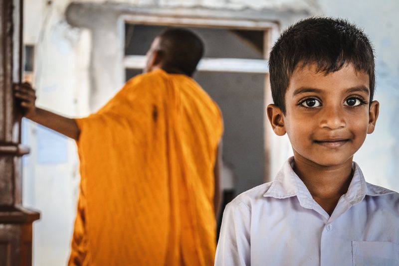 A young boy with huge brown eyes looks directly at the camera. Blurred in the background behind him, a person wearing orange robes can be seen. Taken by photographer Martin Bissig on a Canon EOS R10.
