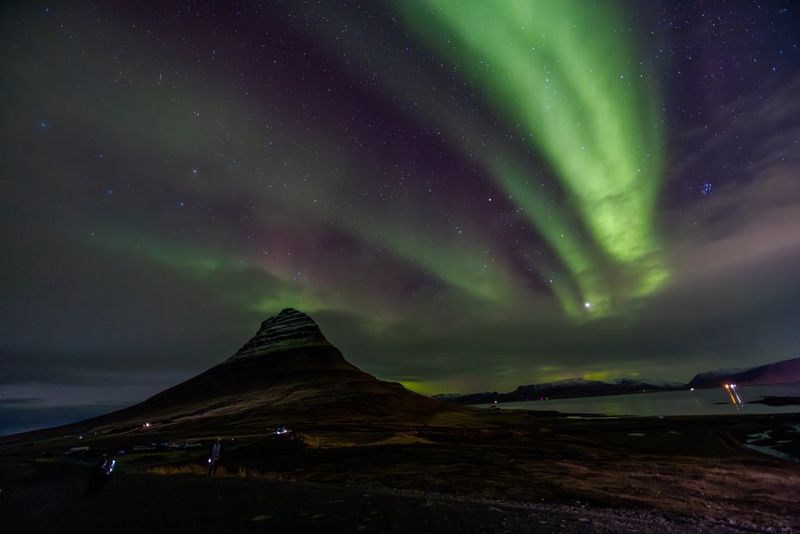 Les aurores boréales apparaissent comme des traînées vertes dans un ciel nocturne sombre au-dessus de la montagne Kirkjufell en Islande, avec quelques personnes et véhicules visibles à sa base. Photo prise par Menna Hossam avec un Canon EOS R5 équipé d'un objectif Canon RF 14-35mm F4L IS USM.