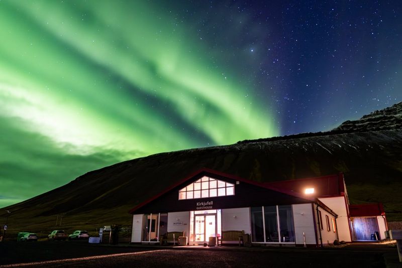 Un bâtiment bas au pied de Kirkjufell est illuminé par l'éclairage artificiel tandis que les aurores boréales se manifestent sous la forme de traînées vertes dans le ciel, la montagne s'élevant derrière. Photo prise avec un Canon EOS R5 équipé d'un objectif Canon RF 14-35mm F4L IS USM. © Menna Hossam