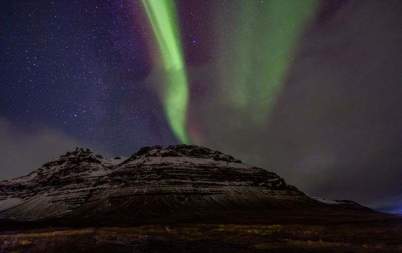 Une traînée verticale d'un vert éclatant se détache sur un ciel sombre et étoilé, au-dessus d'une grande montagne enneigée avec des formations rocheuses en couches distinctes. Photo prise avec un Canon EOS R5 équipé d'un objectif Canon RF 14-35mm F4L IS USM. © Menna Hossam