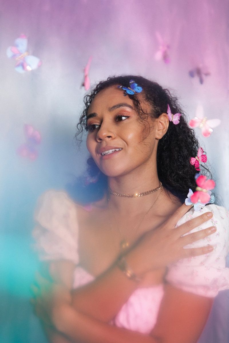 Portrét usmívající se mladé ženy na růžovém pozadí. Hlavu jí obklopují papíroví motýli, kteří zdobí i její vlasy.