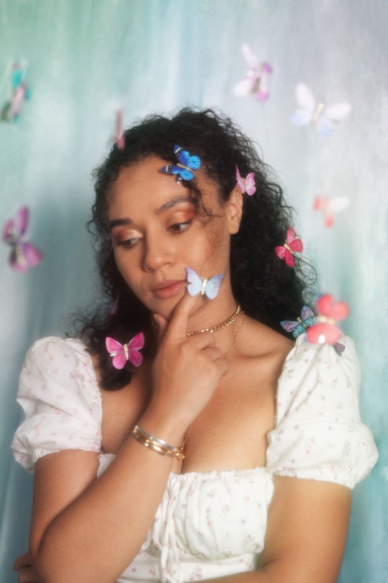 Portrét mladé ženy vystupuje proti zelenému pozadí. Hlavu jí obklopují papíroví motýli, kteří zdobí i její vlasy.