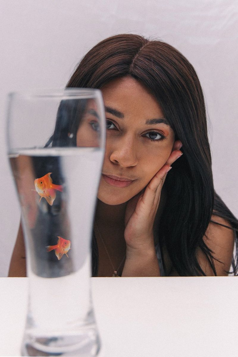Mladá žena naklání hlavu na stranu a opírá se o jednu ruku, zatímco před sebou sleduje sklenici vody, ve které plavou dvě digitálně přidané zlaté rybky.