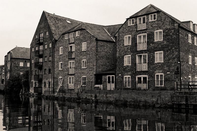 O imagine în nuanţe de sepia a unui şir de depozite de-a lungul unui râu, numeroasele lor geamuri reflectându-se în apa liniştită.