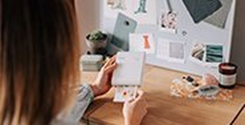 A woman prints out a picture on a Canon SELPHY Square QX10. Positioned upright on the desk in front of her is an outfit inspiration mood board.