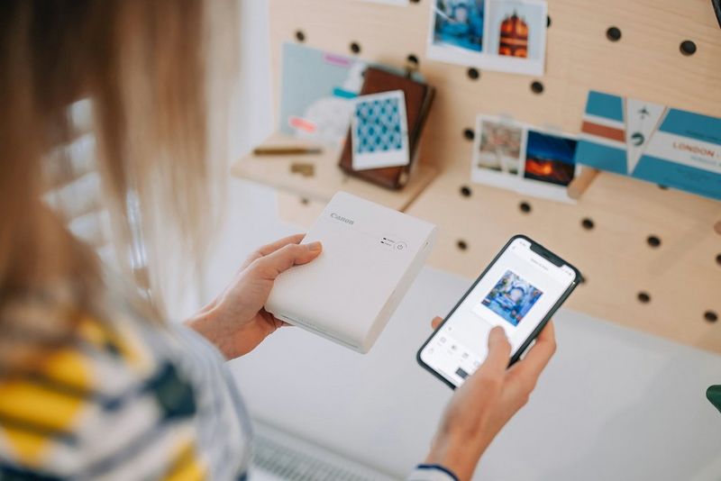 Žena drži pisač SELPHY Square QX10 tvrtke Canon pokraj svog pametnog telefona. Na zaslonu je slika koju se sprema ispisati.
