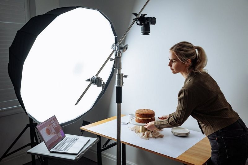 Een vrouw plaatst een taart voor een softboxlamp. Er staat een laptop op een tafel vlakbij en een Canon EOS R8-camera op een statief om van bovenaf te filmen.