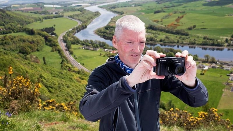 אלן רואן עומד על צד גבעה, המעוטרת בשיחי אולקס צהובים, בשדות מכוסים עשב ונהר בעמק שמאחוריו. הוא אוחז במצלמה קומפקטית כדי לצלם תמונה של הנוף הטבעי.
