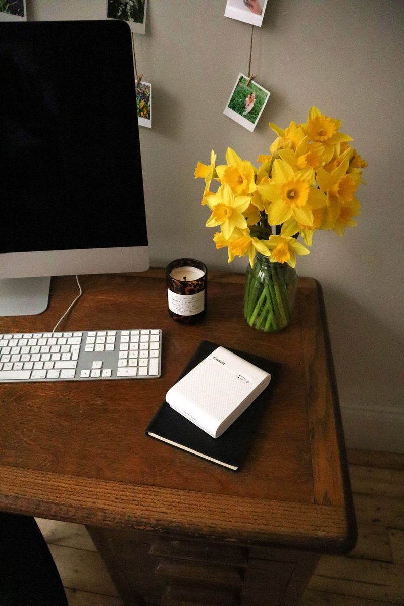 A close-up of a desk containing a computer monitor, keyboard, vase of daffodils and a Canon SELPHY Square QX10 printer.
