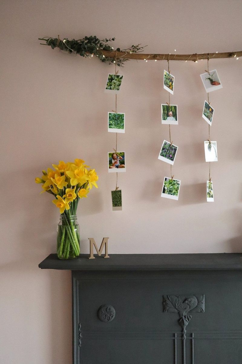 A branch hangs across a wall above a black fireplace. Dropping down from it are several strands of jute with small printed photographs pegged to them.