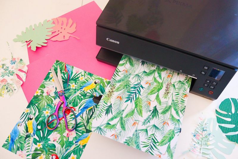 A Canon printer prints out a sheet of paper with a leaf pattern. Next to the printer is another colourful print, as well as several hand-cut paper leaves.