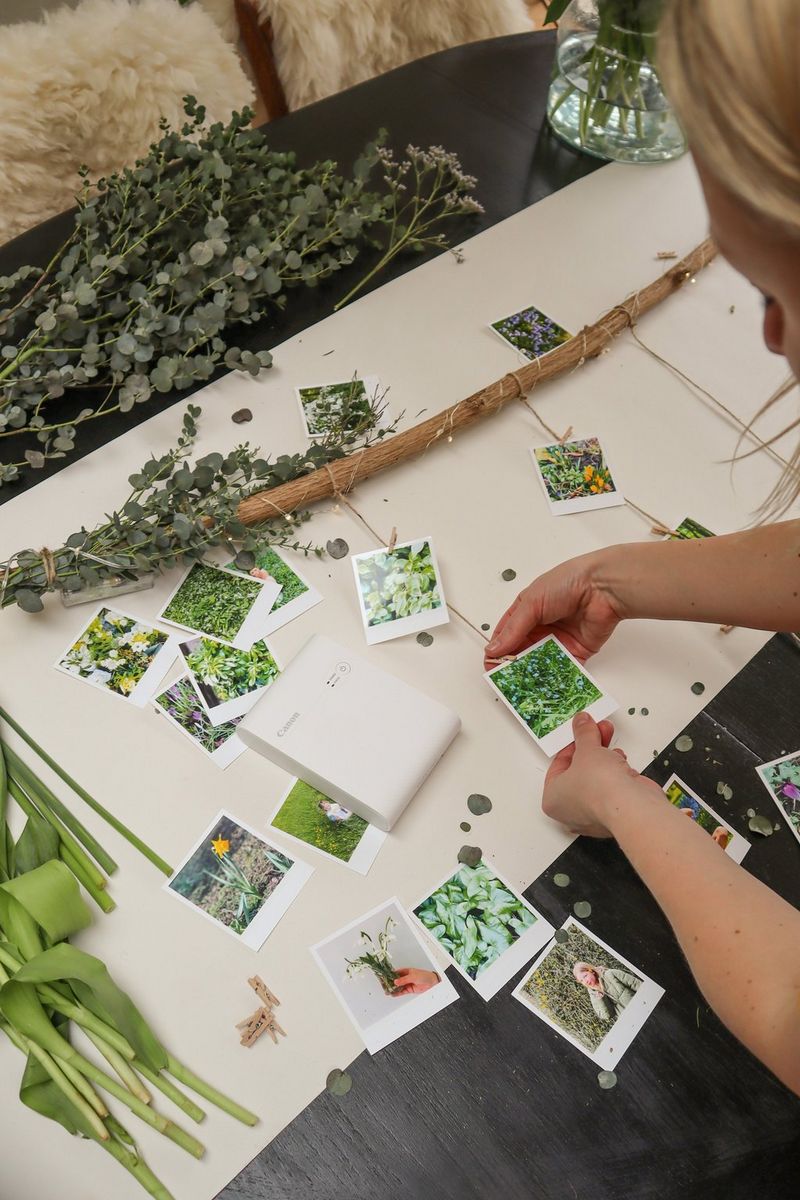 A woman pegs printed photographs of greenery to strands of jute tied to a branch, next to a Canon SELPHY Square QX10 printer.