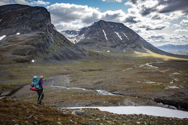 Человек в походном снаряжении, с большим рюкзаком и палками для ходьбы пробирается через горную местность.