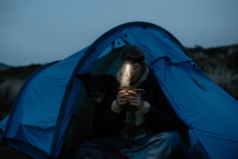 В сумерках человек сидит у палатки, пьет из кружки горячий шоколад, которую освещает луч света от налобного фонарика.