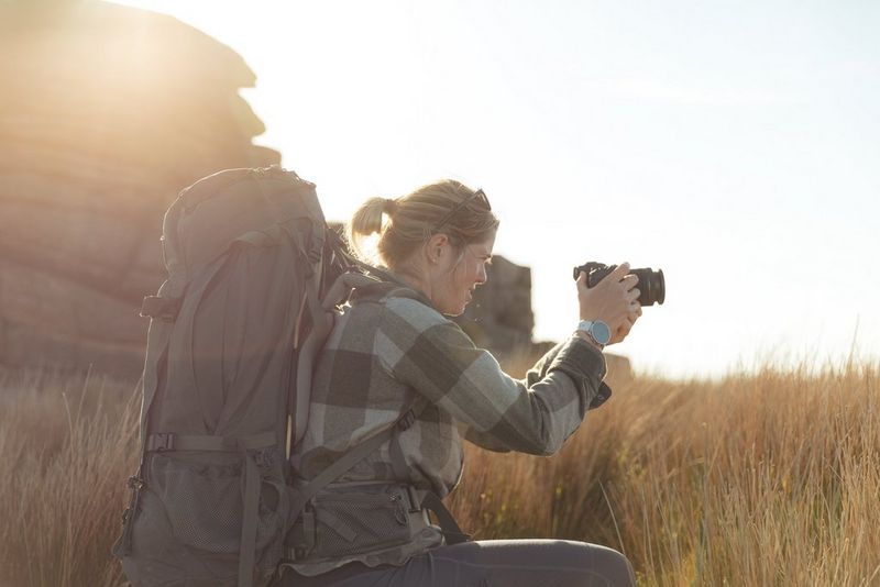 Человек в походном снаряжении и с большим рюкзаком на спине присел в длинной траве, чтобы сделать снимок на камеру Canon.