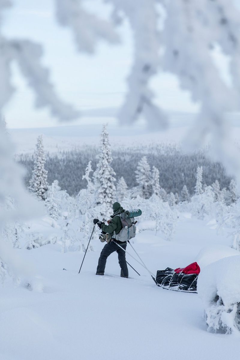 Человек в походном снаряжении тянет за собой сани с вещами и пробирается по глубокому снегу в лесу.