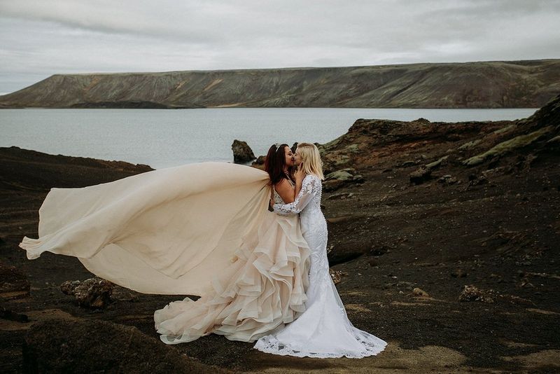 Two female newlyweds kiss on a rocky coastline. The train of one woman's dress billows in the breeze.