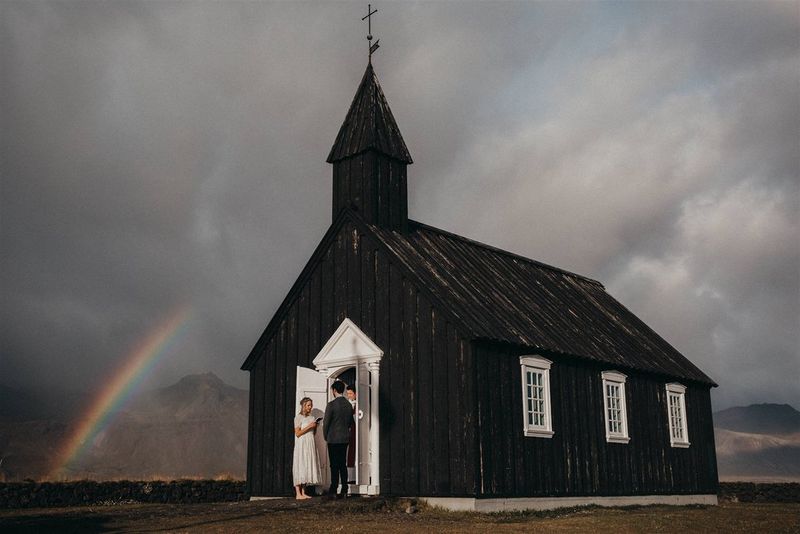 A newlywed couple standing at the white doors of a small black church. A rainbow curves into the grey sky behind them.