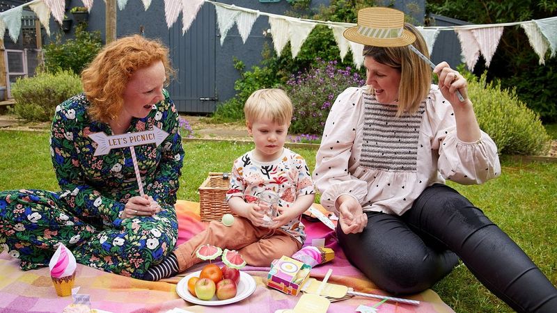 Dvě ženy s malým dítětem uprostřed drží papírové rekvizity do fotostánku a sedí na piknikové dece poseté barevnými papírovými výtvory ze služby Creative Park.
