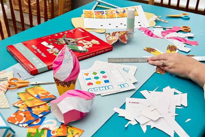 На столе лежат различные печатные объекты, игры и материалы, находящиеся на разных этапах подготовки, а рядом лежит пачка матовой фотобумаги Canon Matte Photo Paper.