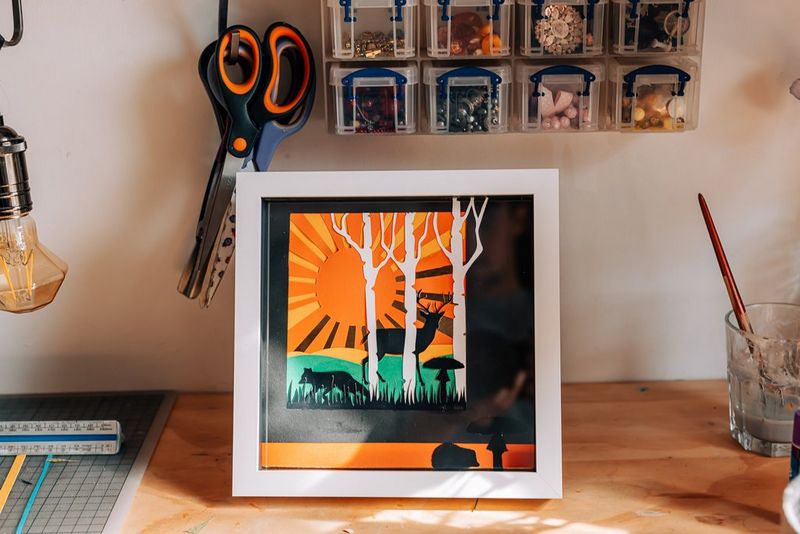 Un paesaggio boschivo realizzato con la carta e incorniciato, posizionato su uno sfondo arancione.