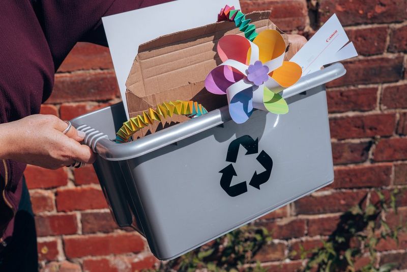 Handen houden een grijze doos vast die is gevuld met stukken karton en papier en weggegooide papierkunstprojecten om te recyclen.