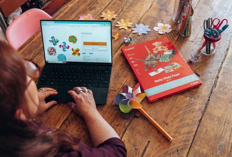 Een vrouw zit achter een laptop met daarop de Canon Creative Park-website. Op de tafel naast haar ligt een pak Canon-printerpapier en staan er een papieren speelgoedwindmolen en potjes met pennen en scharen.