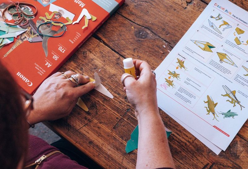 Handen plakken stukken uitgeknipt papier aan elkaar met lijm. Printerpapier en instructies voor het maken van papieren speelgoed liggen ook op tafel.