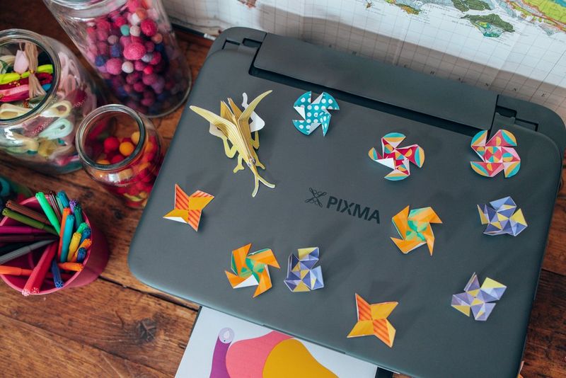 Een selectie kleurrijke papieren speeltjes staat boven op een Canon PIXMA-printer, met pennen en scharen ernaast.