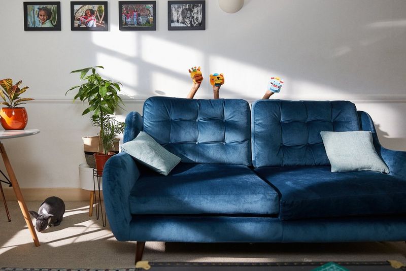 Из-за большого синего дивана в гостиной видны три маленькие ручки с бумажными куклами животных. В левой части кадра виден большой кролик.