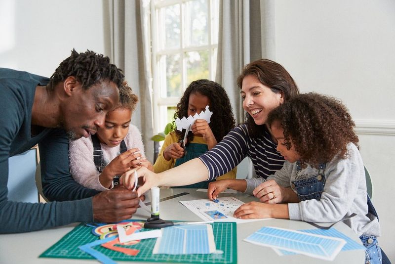 Družinski člani so zbrani za mizo, kjer ustvarjajo nogometno žogo in gol iz papirja.