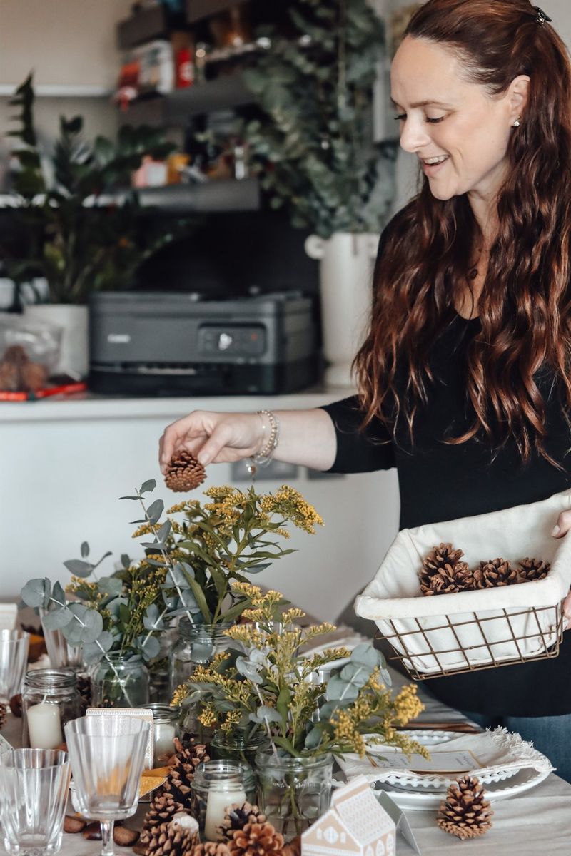 Žena s dlouhými hnědými vlasy pokládá borové šišky po krásně ozdobeném jídelním stole.
