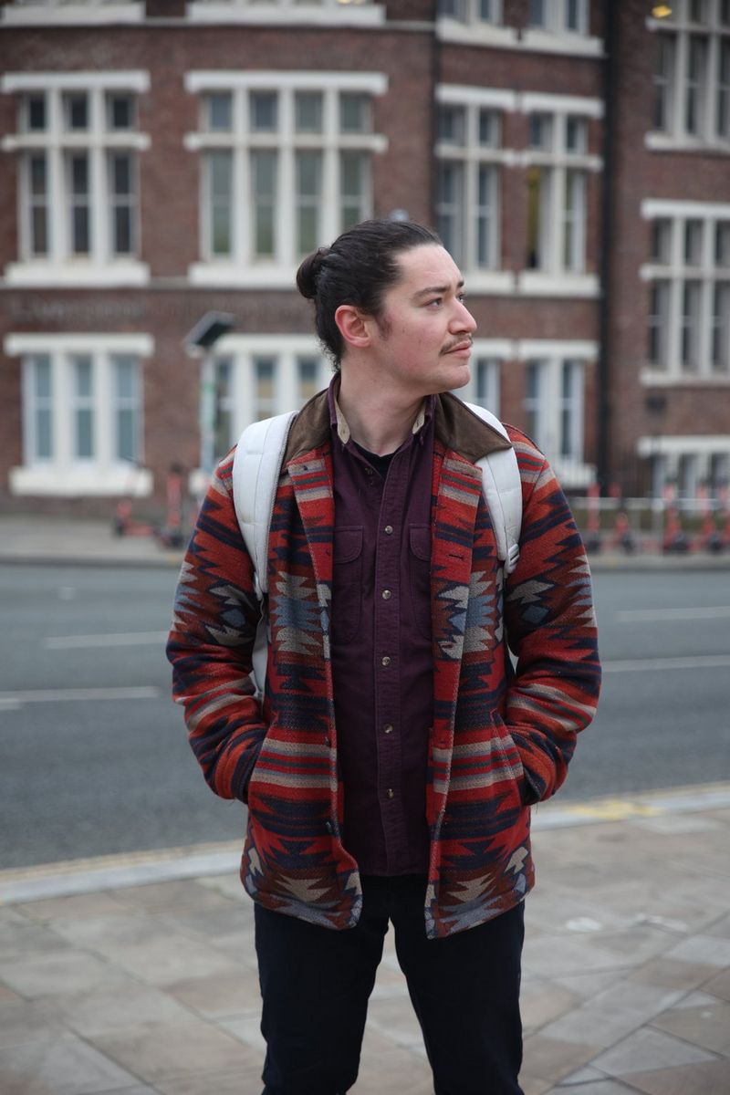 Muškarac u jakni upečatljivog uzorka, s tamnom kosom vezanom u punđu, stoji s rukama u džepovima kaputa, a glava mu je okrenuta u stranu.