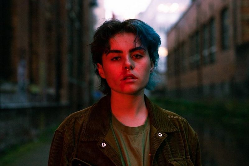 Oseba s kratkimi obarvanimi lasmi in obročastim uhanom v nosu gleda proti fotoaparatu.