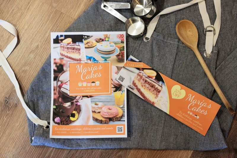 Odštampani poster i baner koji promovišu poslastičarnicu postavljeni na sivu kecelju zajedno sa šoljama za merenje i drvenom kašikom.