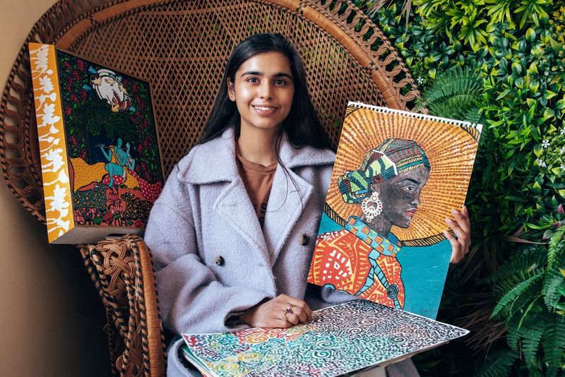 A young woman sitting in a wicker chair proudly holds up a painting. On her lap and next to her are two more colourful artworks. 