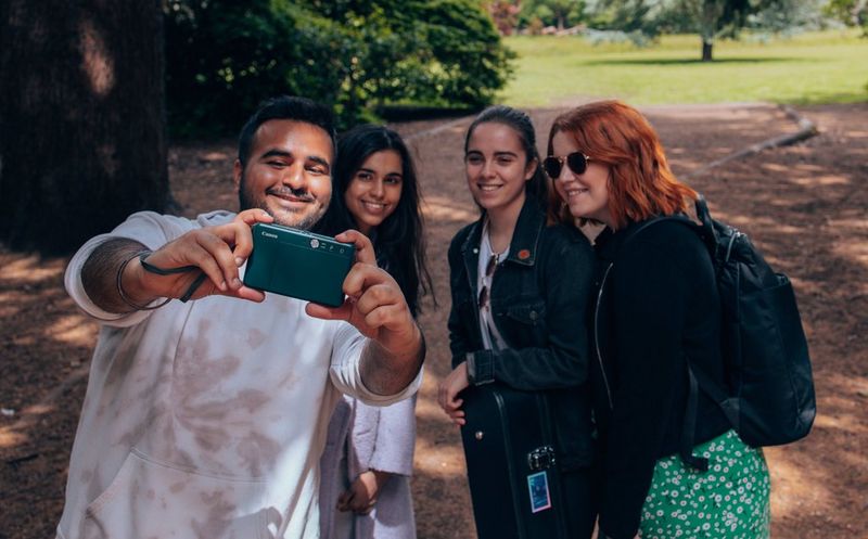 Four young people pose for a group selfie in a park, being taken with a Canon printer camera.