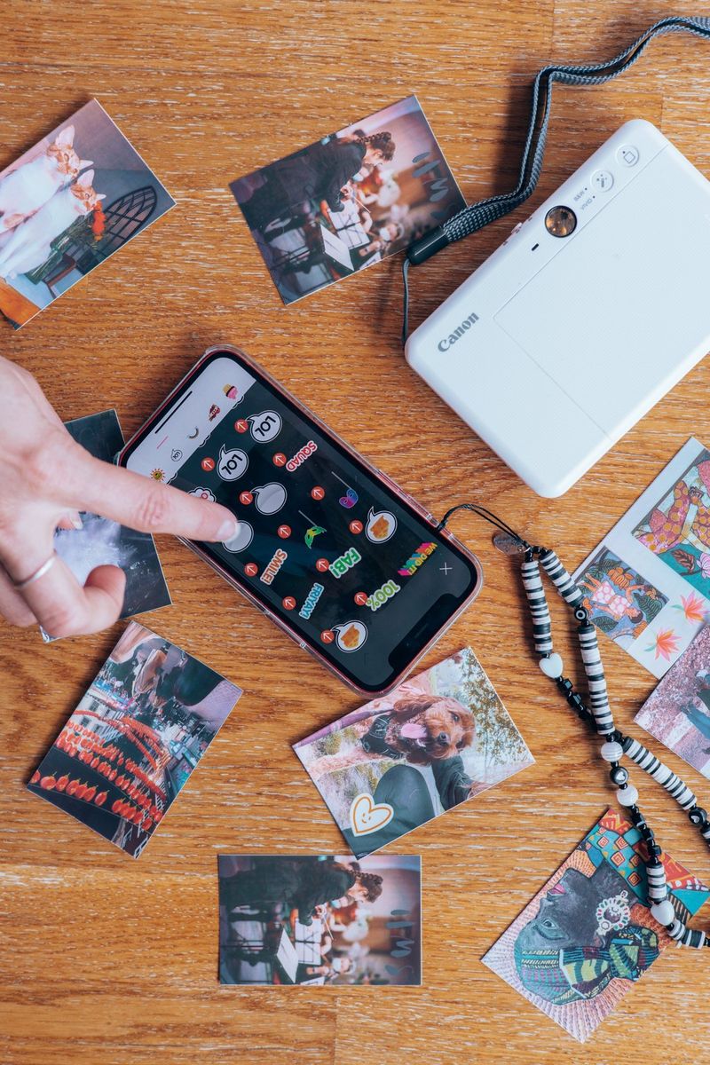 A Canon Zoemini S2, smartphone and selection of prints are laid out on a wooden surface. A hand can be seen using the touchscreen on the smartphone.