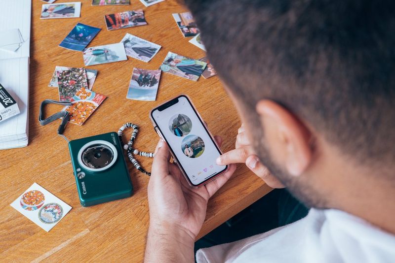 A person using the Canon Mini Print app on a smartphone to make their photos into circular sticker prints. The table in the background is covered with Canon Zoemini S2 print outs.
