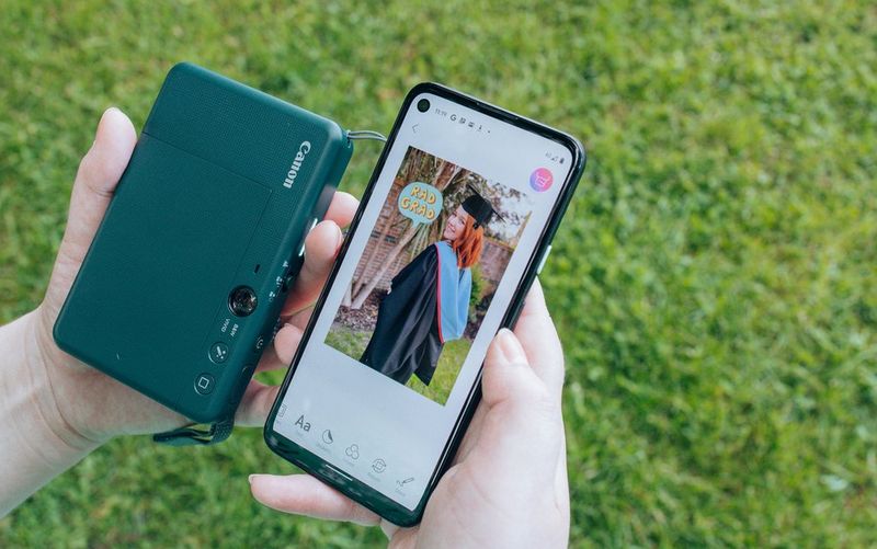 A pair of hands hold up a Canon Zoemini S2 and a smartphone showing the Canon Mini Print app, with a graduation photo on its screen.