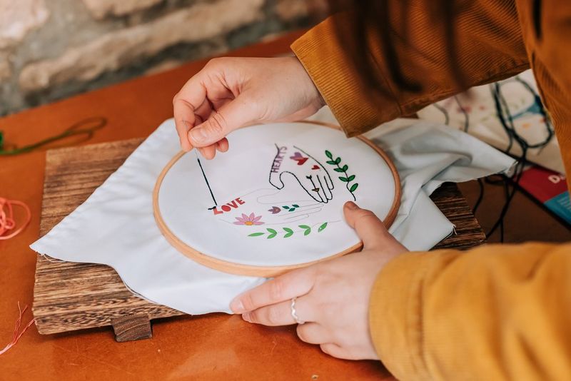 Женщина пришивает узор в виде рук по контуру к куску ткани.