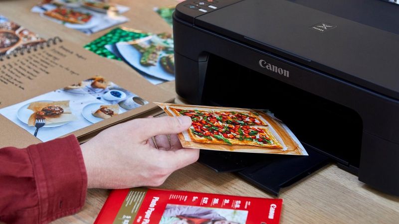 A hand taking a print of an image of a sweet potato tart from the tray of a PIXMA printer, alongside a pack of printer paper and a homemade recipe book.