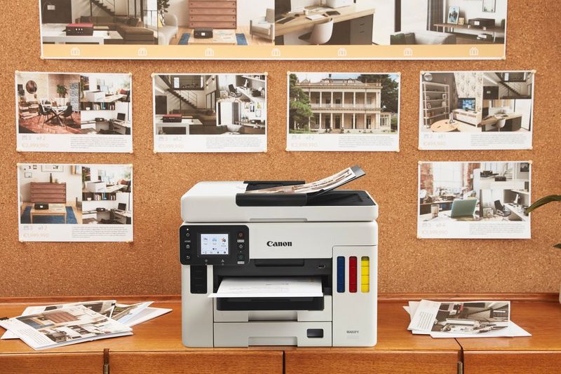 Een MAXIFY GX6050-printer met navulbare inkt, met aan de voorzijde kijkvensters voor het inktniveau en prints die op een mededelingenbord erachter zijn vastgeprikt.